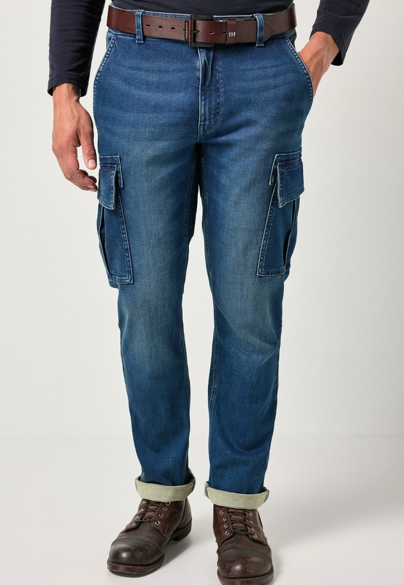 Man in opgerolde blauwe spijker cargo shorts, bruine leren riem, donker overhemd met lange mouwen en bruine leren veterschoenen staat.