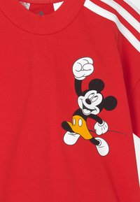 Czerwona koszula z prążkowanym kołnierzykiem i białymi paskami na ramionach, przedstawiająca uśmiechającego się Myszka Mickey w żółtych szortach i białych rękawicach, podnoszącego pięść.