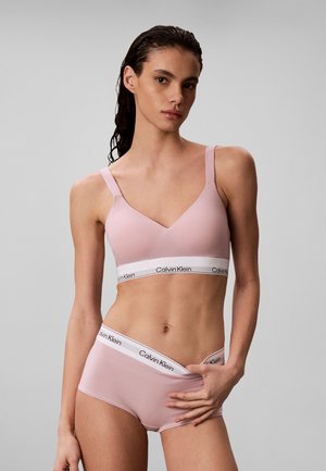 Kobieta nosząca jasnoróżowy biustonosz Calvin Klein i pasującą bieliznę z markowymi elastycznymi taśmami, stojąca na jednolitym szarym tle.