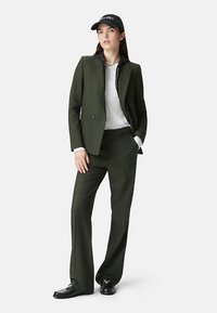 Blazer y pantalones verde oscuro a medida, camisa blanca, gorra negra con texto "CAMERA", zapatos negros, corte slim, tela lisa, diseño estructurado.