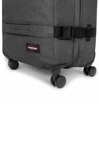 Grauer Stoffkoffer mit schwarzem Besatz, versehen mit einer Schnallenschließe, einem Markenpatch und vier Rollen für Mobilität.