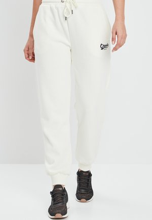Witte sweatpants met elastische boorden, een trekkoord in de taille en een zwart logopatch op de linkerheup. Gecombineerd met zwart gemusterde sneakers.