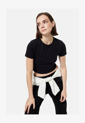 Ung kvinde i sort crop top og sorte bukser med hvid sweatshirt bundet om livet, kigger til siden mod en ensfarvet baggrund.