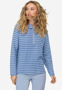 Długi rękaw, niebieski sweter w paski z pół-zamek i kołnierzykiem. Wykonany z lekkiego materiału, z beżowymi paskami, noszony z jasnoniebieskimi spodniami.