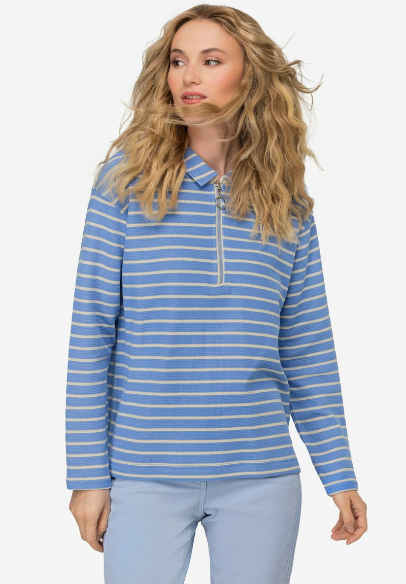 Długi rękaw, niebieski sweter w paski z pół-zamek i kołnierzykiem. Wykonany z lekkiego materiału, z beżowymi paskami, noszony z jasnoniebieskimi spodniami.