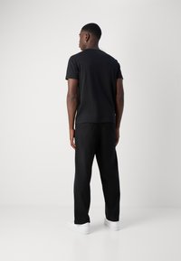 Svart t-skjorte med korte ermer, brukt med svarte bukser og hvite sneakers, sett fra baksiden mot en ensfarget lys bakgrunn.