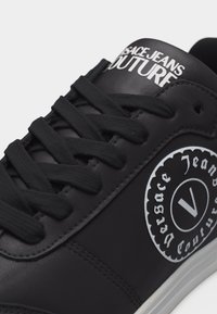 Μαύρο αθλητικό παπούτσι Versace Jeans Couture με μαύρα κορδόνια και λευκό λογότυπο στην πλευρά και στη γλώσσα, με λευκή σόλα.