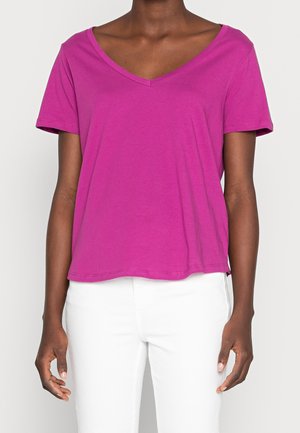 Femme portant un haut magenta à manches courtes et col en V, ainsi qu'un pantalon blanc, vue des épaules jusqu'aux hanches sur un fond uni.