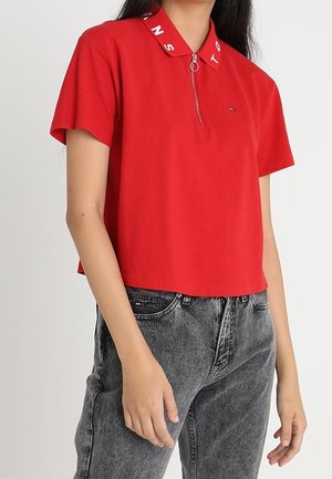 Femme portant un polo rouge à manches courtes avec un col zippé et un jean noir délavé, debout, les mains relâchées le long du corps.