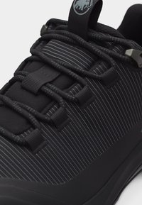 Prim-plan cu un pantof sport negru cu model reflectorizant cu dungi, șireturi groase negre și un șnur de tragere care prezintă un logo cu un mamut.