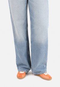 Jeans de mezclilla anchos de color azul claro combinados con zapatillas naranjas con cordones blancos y una suela de goma marrón.