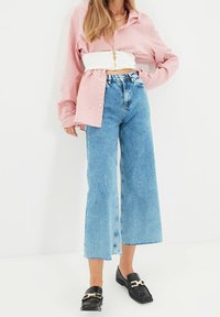 Nainen, jolla on päällään vaaleanpunainen oversized-paita valkoisella lyhyellä vetoketjyouksityiskohdalla, korkeavyötäröiset leveät siniset farkut ja mustat loafertsit kultaisilla soljilla.