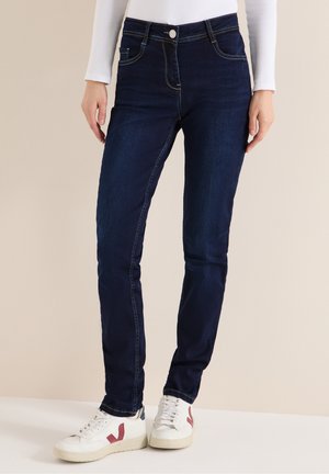 Personne portant un jean slim bleu foncé et des baskets blanches avec un logo rouge, debout sur un fond neutre.