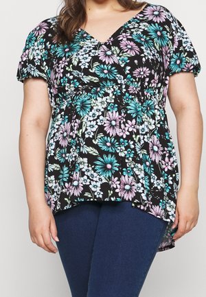 Frau trägt eine schwarze Bluse mit kurzen Ärmeln und V-Ausschnitt, verziert mit türkis-, lavendel- und weißen Blumen, kombiniert mit dunkelblauen Jeans.