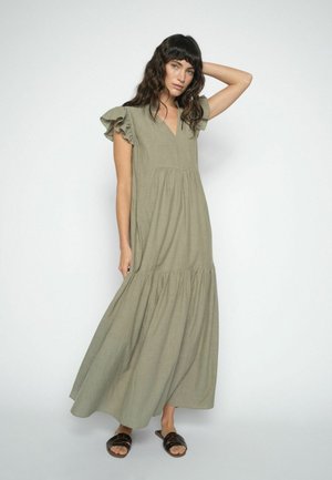 Kvinde med langt mørkt hår iført en olivengrøn maxikjole med flæset ærme og sorte slidesandaler foran en ensartet hvid baggrund.