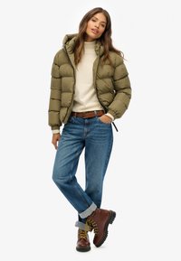 Olijfgroene pufferjack met capuchon, crèmekleurige trui, blauwe spijkerbroek en bruine veterschoenen met gele accenten. Casual winteroutfit.