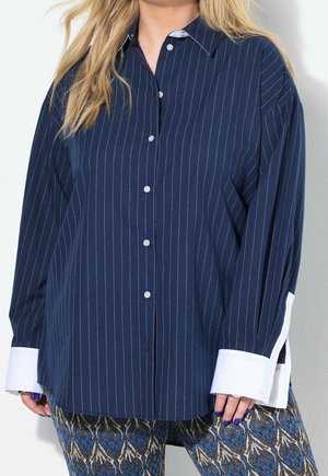 Femme portant une chemise boutonnée à fines rayures bleu marine avec des poignets blancs surdimensionnés et un pantalon bleu et beige à motifs.