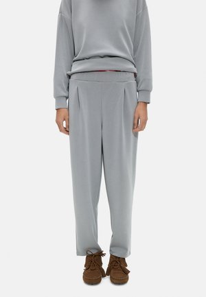 Osoba nosząca jasnoszare plisowane spodnie i pasującą bluzkę z długim rękawem, zestawione z brązowymi butami w stylu mokasynów.