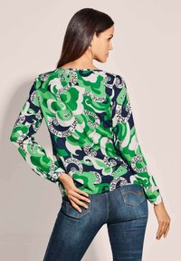 Langärmlige Bluse mit einem grün-blauen Muster, das wirbelnde Designs und Städtenamen enthält, aus einem glatten, leichten Stoff gefertigt.