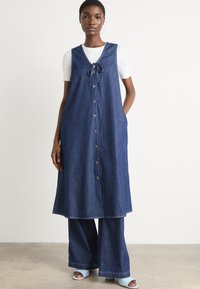 Mørkeblå denimkjole med knytehals og knappestolpe. Brukt over en hvit t-skjorte med vide denimbukser og åpne sko.