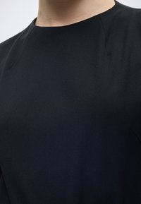 Κοντινό πλάνο του άνω μέρους του στήθους και του λαιμού που φοράει ένα απλό μαύρο τοπ με στρογγυλή λαιμόκοψη και διακριτικές ραφές κοντά στους ώμους.