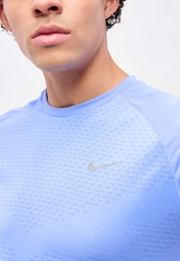 Junge Person trägt ein hellblaues Nike-Sportshirt mit strukturiertem Muster und reflektierendem Swoosh-Logo auf der Brust.