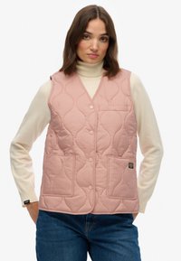 Gilet matelassé rose clair avec col montant, boutons-pression et deux poches avant. Porté sur un pull à col roulé crème, avec un détail d'étiquette.