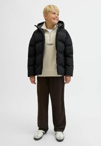 Lys-haired dreng smilende, iført en sort pufferjakke, beige lynlåset sweatshirt, mørke bukser og hvide sneakers med sorte detaljer, stående.