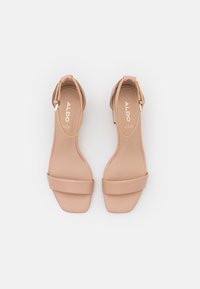 Nude high-heeled sandals made of smooth synthetic material. Features an ankle strap, square toe, and a flat heel with gold-tone accents.