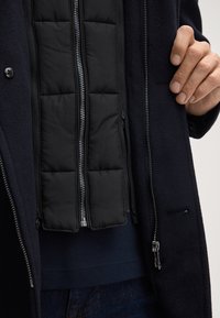 Giacca blu scuro con un interno trapuntato nera con zip. Caratteristiche includono un tessuto esterno testurizzato, più cerniere e un design aderente.