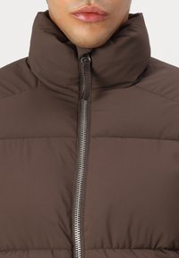 Hnědá péřová bunda s vysokým límcem a zapínáním na celý zip. Materiál je hladký s prošívanou izolací pro tepelný komfort.