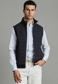 Vesta matlasată de culoare navy, cu guler înalt, fermoar frontal și tiv elastic, purtată peste o cămașă cu nasturi de culoare albastru deschis, asortată cu pantaloni bej.