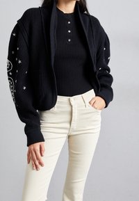 Gilet en tricot noir avec une texture côtelée, featuring un col châle et des motifs d'étoiles sur les manches, associé à un haut noir ajusté et un pantalon clair.