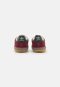 Røde semskede sneakers med en grøn hælflap, gennemsigtig gummisål og hvid Adidas-logo på hælen. Bagfra.