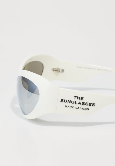 Valged prillid, millel on ümbritsev disain, halli tooni objektiivid ja pintseldatud tekst "THE SUNGLASSES MARC JACOBS" kaanel.