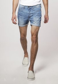 Pantalones cortos de mezclilla de color azul claro con dobladillo enrollado, diseño de cinco bolsillos y cierre de botón metálico, combinados con zapatos blancos tipo slip-on.