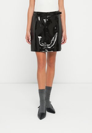 Femme portant une mini-jupe en vinyle noir brillant avec une ceinture assortie, associée à des chaussettes grises et à des talons pointus noirs, debout sur un fond blanc.