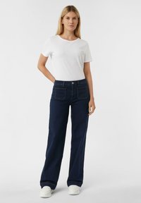 Jeans a gamba larga di un blu scuro con tasche frontali e posteriori, abbinati a una maglietta bianca semplice e a delle sneakers bianche. Tessuto di cotone liscio.