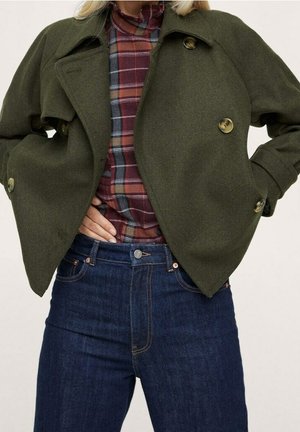 Frau trägt eine olivgrüne, mit Knöpfen geschlossene Jacke über einem rot karierten Hemd und dunkelblaue hoch taillierte Jeans, die Hände in den Taschen.