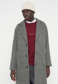 Pilkas zigzago rašto paltas su atlapais ir juodais mygtukais, dėvimas virš bordo spalvos džemperio su auksinėmis siuvinėtomis detalėmis. Smėlio spalvos megzta kepuraitė.