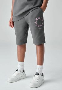 Osoba oblečená v sivých šortkách s ružovým logom "ALLSAINTS LONDON", biele ponožky "ALLSAINTS" a biele tenisky, stojaca s jednou rukou v vrecku.