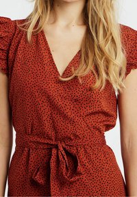 Červené šaty áčkového střihu s puntíky, krátkými nabíranými rukávy, hlubokým V výstřihem a tkanicemi v pase. Lehký a strukturovaný materiál.