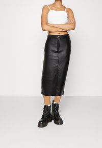 Kvinna med korslagda armar som bär en vit axelbandslös crop top, en svart högmidjad läder midi-kjol och svarta snörstövlar med vita strumpor.