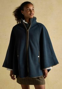 Žena v tmavomodrom plášti s vysokým golierom a zipsom, skombinovanom s kockovanou sukňou, stojaca na béžovom pozadí.