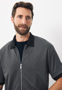 Mand med brunt krøllet hår og skæg iført en kortærmet grå mønstret zip-up skjorte med sort krave, som kigger let til siden.