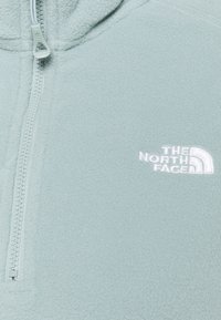 Pullover in pile di colore azzurro chiaro con zip frontale. Presenta logo ricamato bianco sul petto e superficie in tessuto texturizzato.
