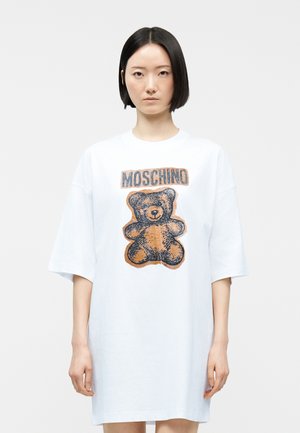 Nő rövid fekete hajjal, visel egy túlméretezett fehér pólóruhát, amelyen barna maci és "MOSCHINO" felirat látható elöl.