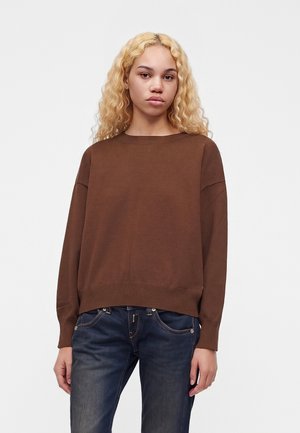 Giovane con capelli biondi ricci che indossa un maglione marrone a maniche lunghe e jeans blu scuro, in piedi su uno sfondo semplice.
