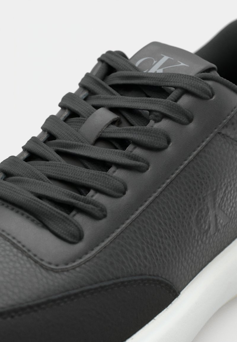 Zwarte leren sneaker met gestructureerde panelen, platte zwarte veters en een grijs logo op de tong. Witte rubberen zool aan de onderkant.