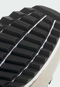 Sulan av en sneaker med svart gummi med texturerade, upphöjda diamantformer, en vit mellansula och beige sidopaneler.